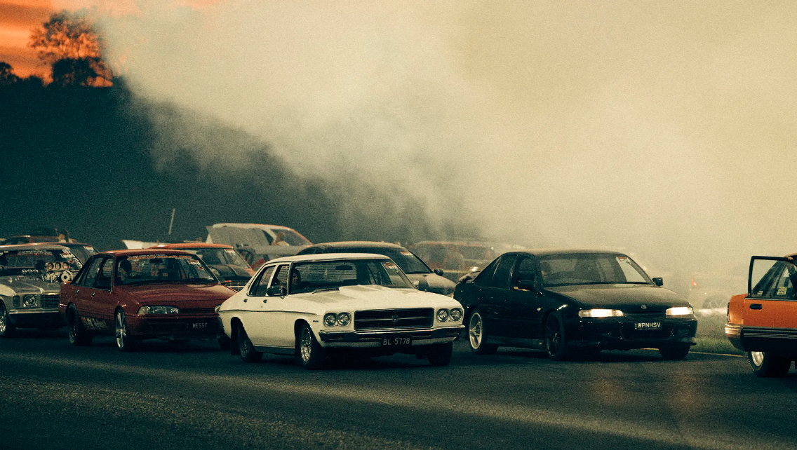 Several classic cars line up on a track, smoke partially obscuring the view. Racing cars are staged on the track for a vintage car event.