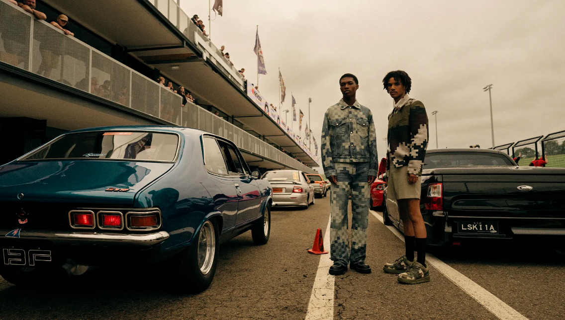 Two men posing with a blue vintage car and black truck at a racetrack with spectators, creating a fashion editorial scene.