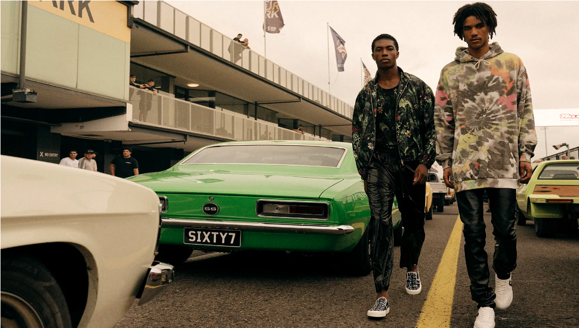 Two men model fashion streetwear with classic cars on a racetrack; one in a floral jacket, the other in a tie-dye hoodie.