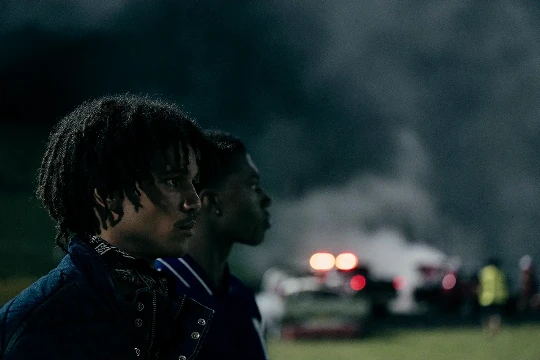 Two young Black men in a dimly lit outdoor scene with smoke and blurred red lights in the background, captured in a medium shot.