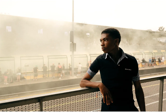 Portrait of a young adult Black man leaning on a railing, looking off to the side with people and smoky haze in the background.