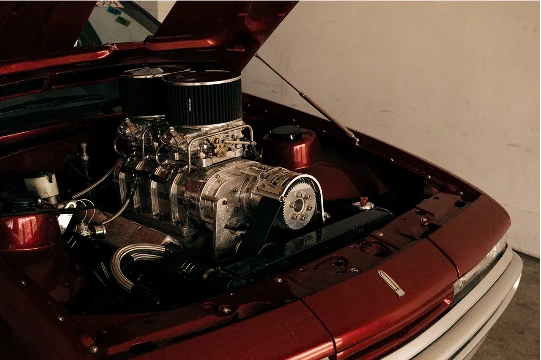 Open hood of a red car reveals a complex chrome engine with black air filters, set against a neutral-toned indoor background.