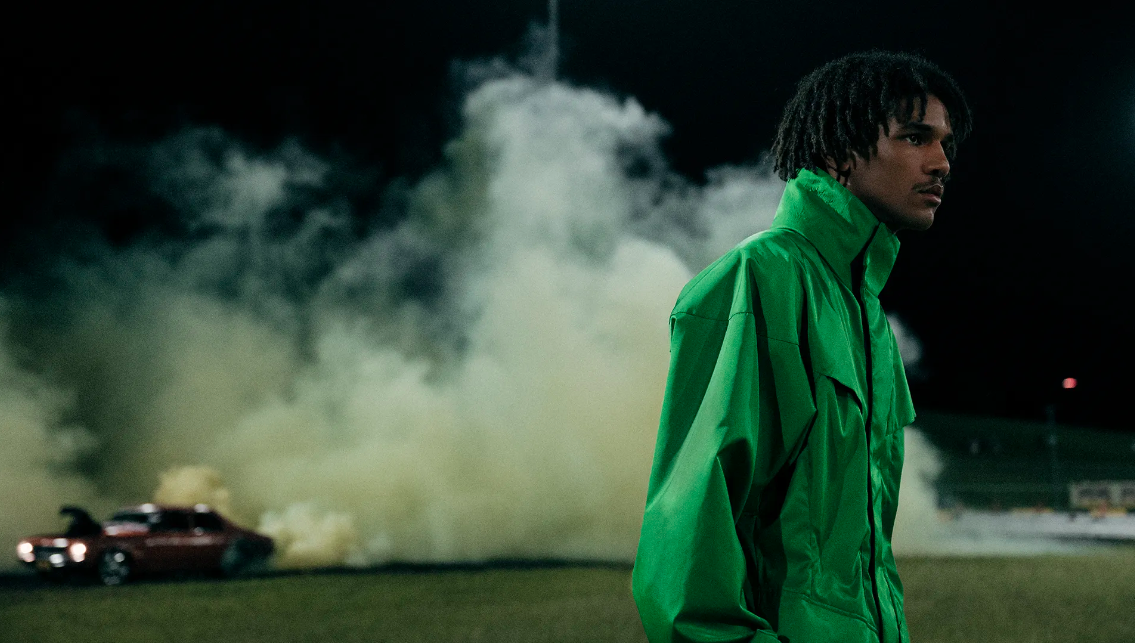 Portrait of a handsome young adult male in a green jacket standing on grass at night, with a vintage car and smoke in the background.