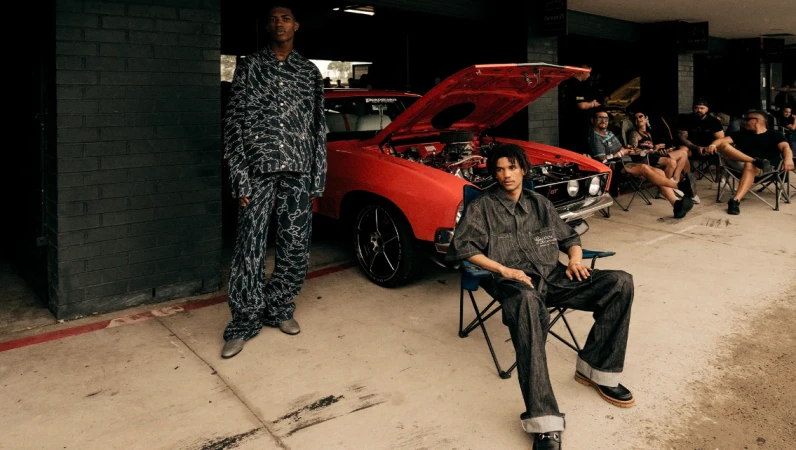 Two adult males pose next to a vintage red muscle car with an open hood at a garage gathering with other adults in the background.