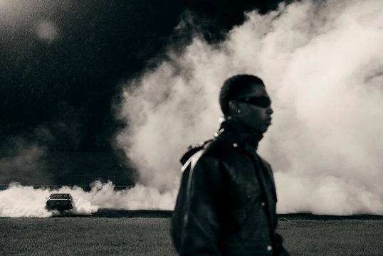 Monochrome portrait of a person in a leather jacket against smoke, with a car visible in the background on a field at night.