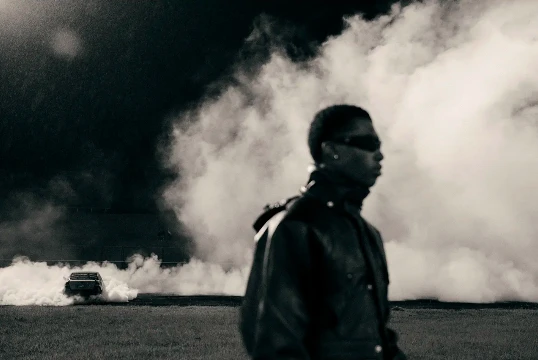 Monochrome portrait of a person in a leather jacket against smoke, with a car visible in the background on a field at night.