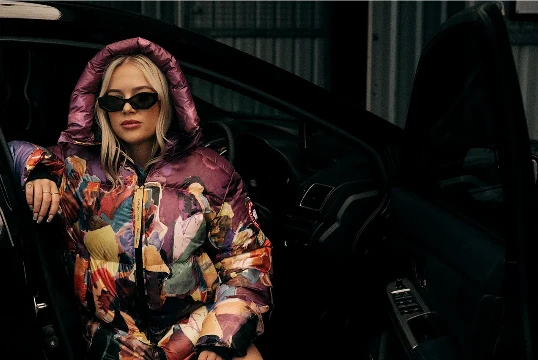 Portrait of a young, blond woman wearing black sunglasses and a patterned puffer jacket, sitting in the driver's seat of a car.