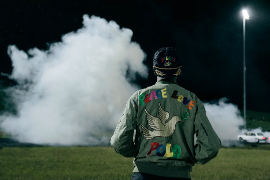 Back view of a man in a decorated olive-green jacket and baseball cap against a dark sky with bright vapor and cars at night.