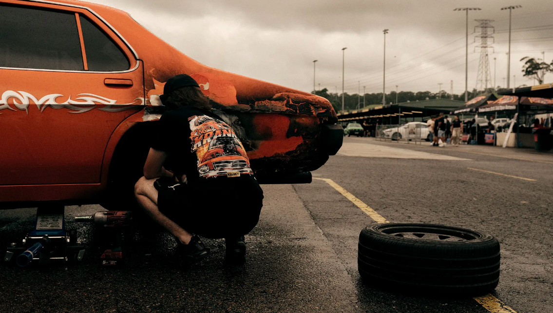 A man crouches near an orange classic car in an outdoor setting, working on the tire, with tools and a spare tire visible nearby.