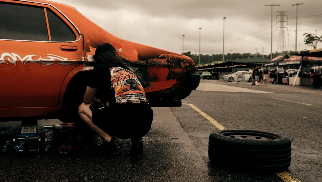 A man crouches near an orange classic car in an outdoor setting, working on the tire, with tools and a spare tire visible nearby.