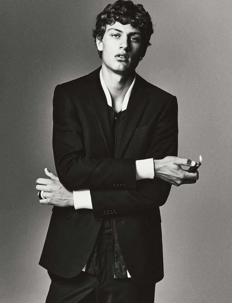 Monochrome portrait of a curly-haired man in a black suit with his arms crossed while holding a lighter in an indoor setting.