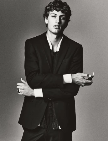 Monochrome portrait of a curly-haired man in a black suit with his arms crossed while holding a lighter in an indoor setting.