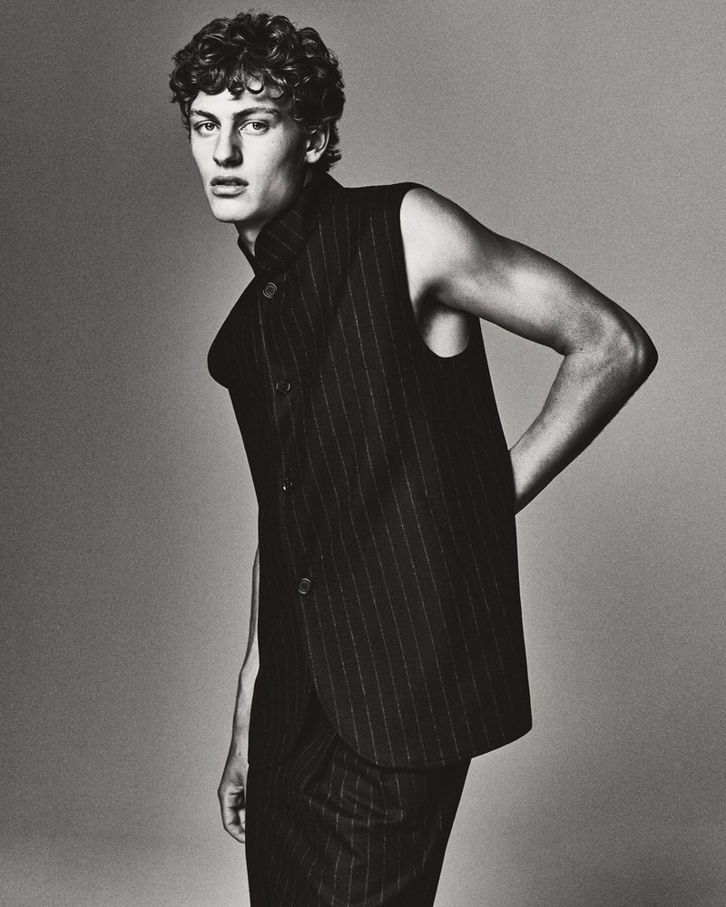 Monochrome portrait of a young adult male with curly hair, wearing a pinstripe vest and pants, posing in front of a neutral background.