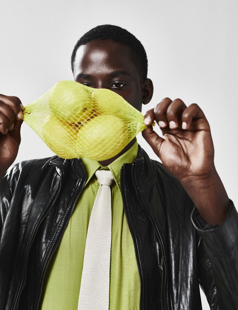 Dark-skinned man in black leather jacket and light green shirt poses holding yellow mesh bag with lemons over lower face, studio lighting.