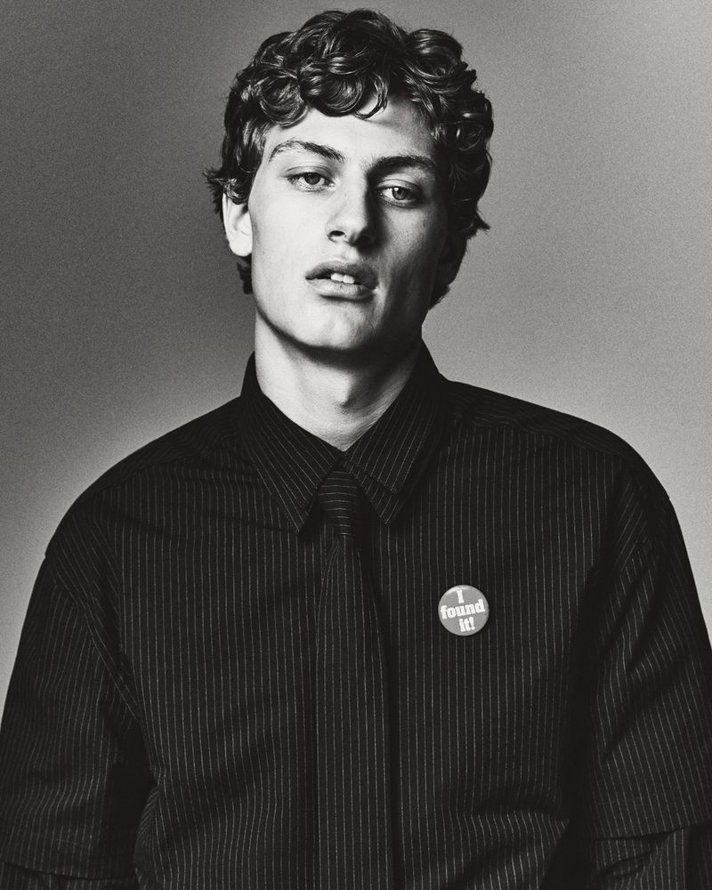 Monochrome portrait of a young adult male with curly hair, wearing a striped shirt, against a plain grey backdrop, emphasizing a classic studio aesthetic.
