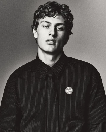 Monochrome portrait of a young adult male with curly hair, wearing a striped shirt, against a plain grey backdrop, emphasizing a classic studio aesthetic.