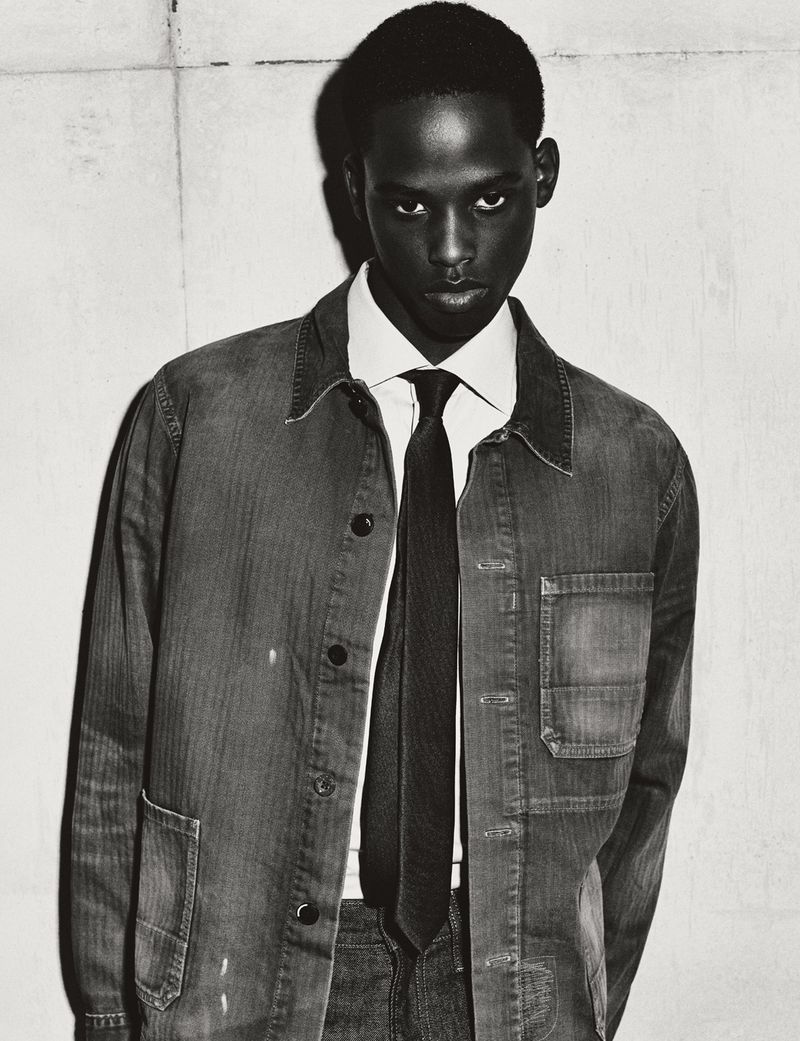 Portrait of a black man wearing a collared shirt, tie, and denim jacket against a plain wall.