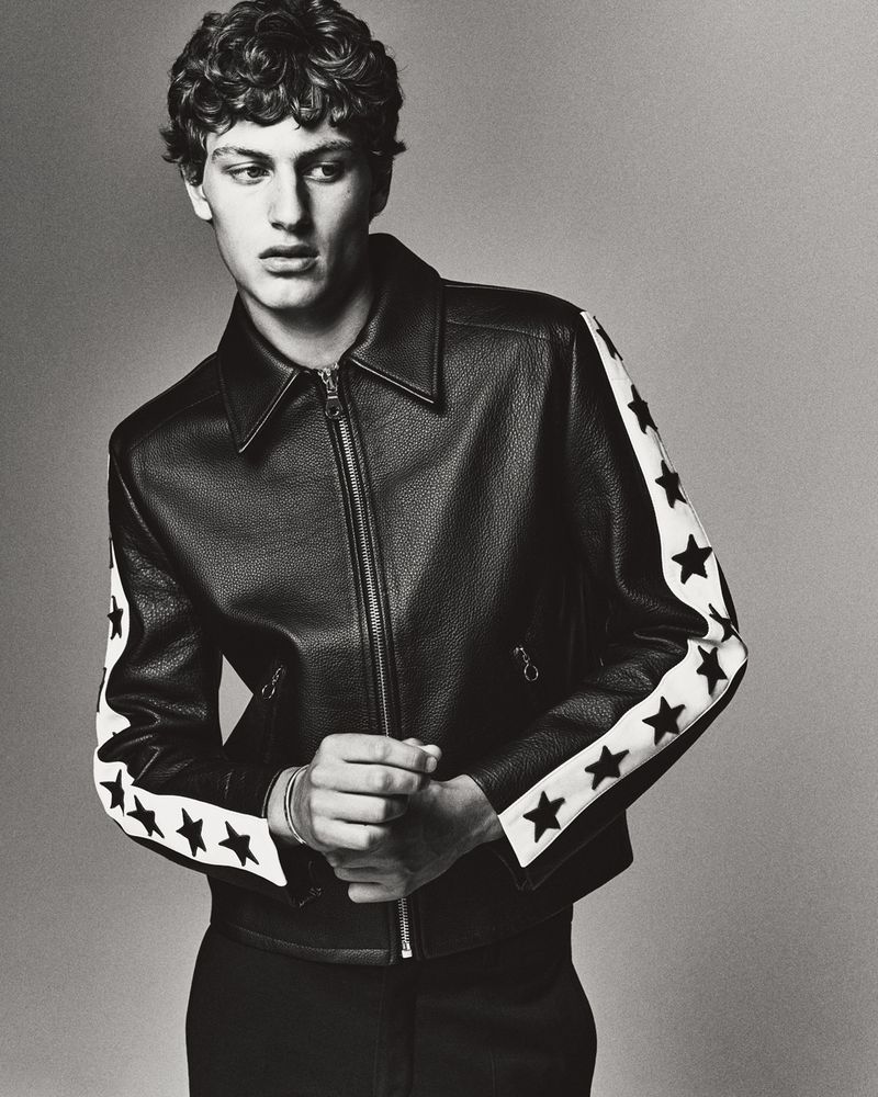 Monochrome portrait of a young adult man with curly hair wearing a black leather jacket with white star-patterned sleeves.