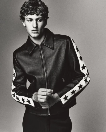 Monochrome portrait of a young adult man with curly hair wearing a black leather jacket with white star-patterned sleeves.