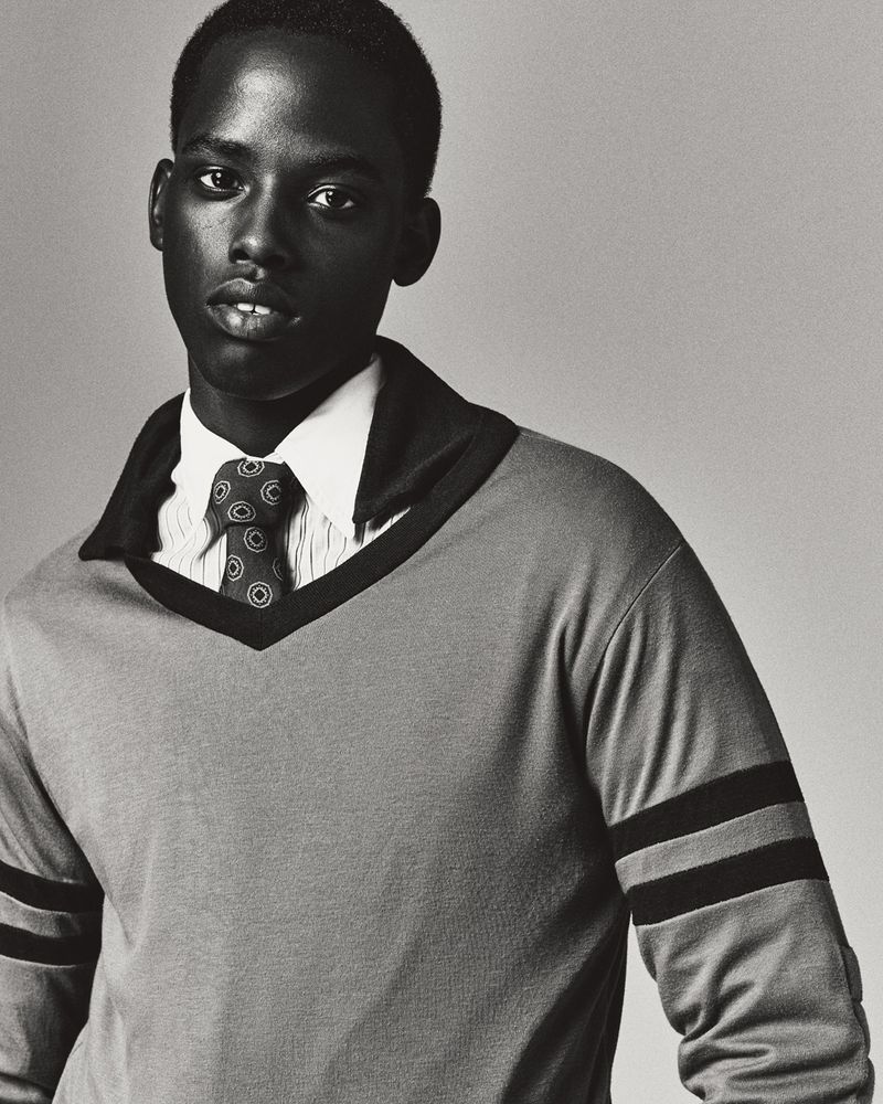 Monochrome portrait of a young Black man in a collared sweater and tie, posing indoors against a plain background in a close-up shot.