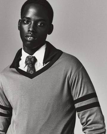 Monochrome portrait of a young Black man in a collared sweater and tie, posing indoors against a plain background in a close-up shot.