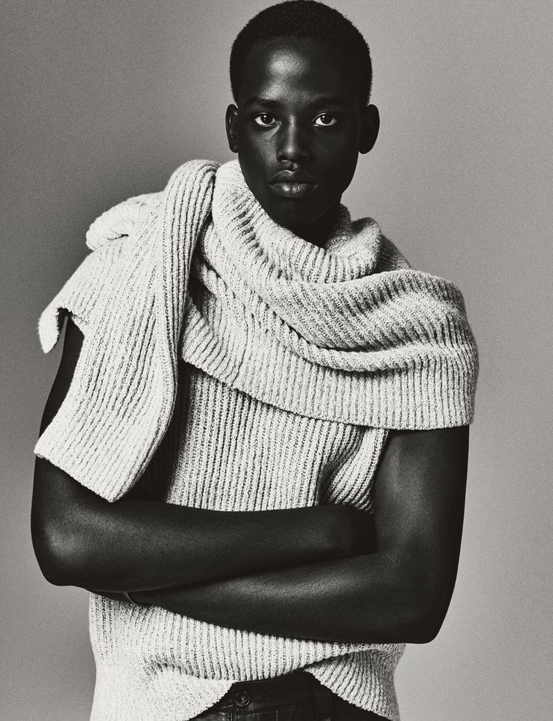 Portrait of a Black man with short hair wearing a cream-colored ribbed knit sweater with a scarf draped over his shoulder against a grey background.