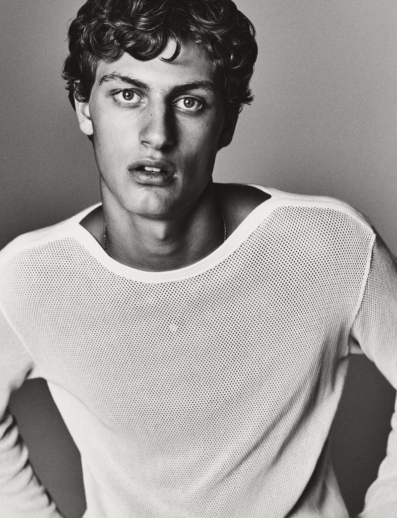 Portrait of a young adult man with curly hair, wearing a white top, gazing at the camera against a neutral-toned background in a photo studio.