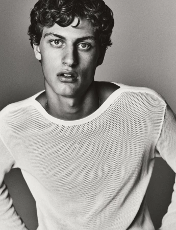 Portrait of a young adult man with curly hair, wearing a white top, gazing at the camera against a neutral-toned background in a photo studio.