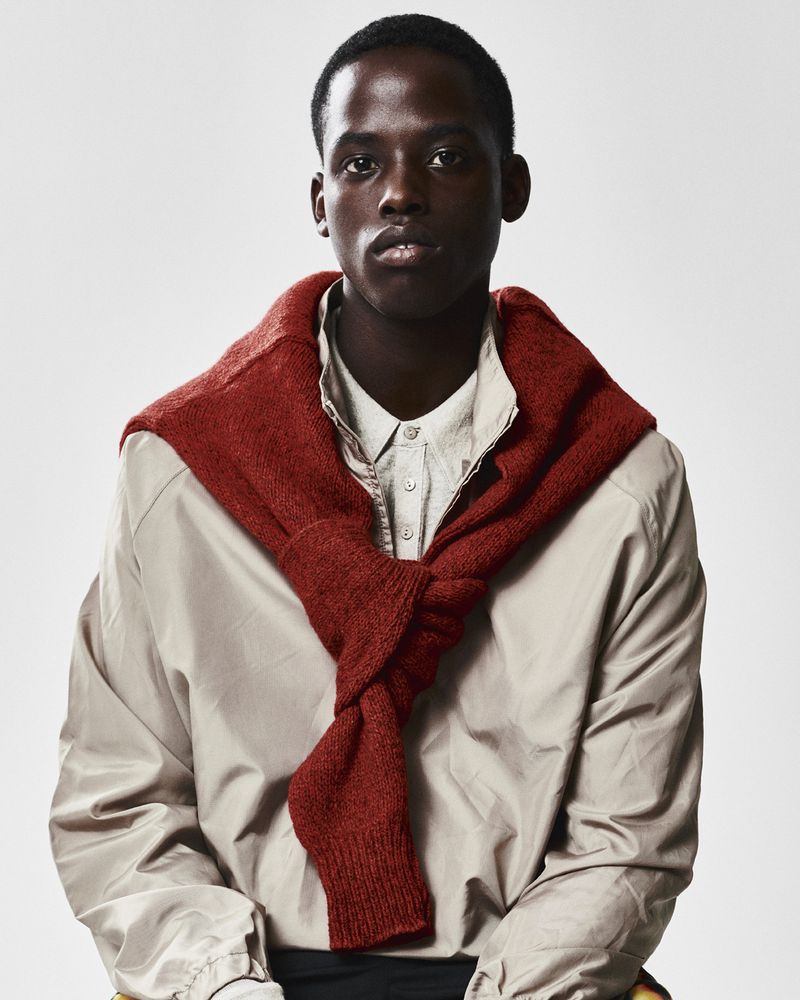 Portrait of an African American man in a casual outfit: light jacket, button-down shirt, and rust-colored sweater draped around his shoulders.