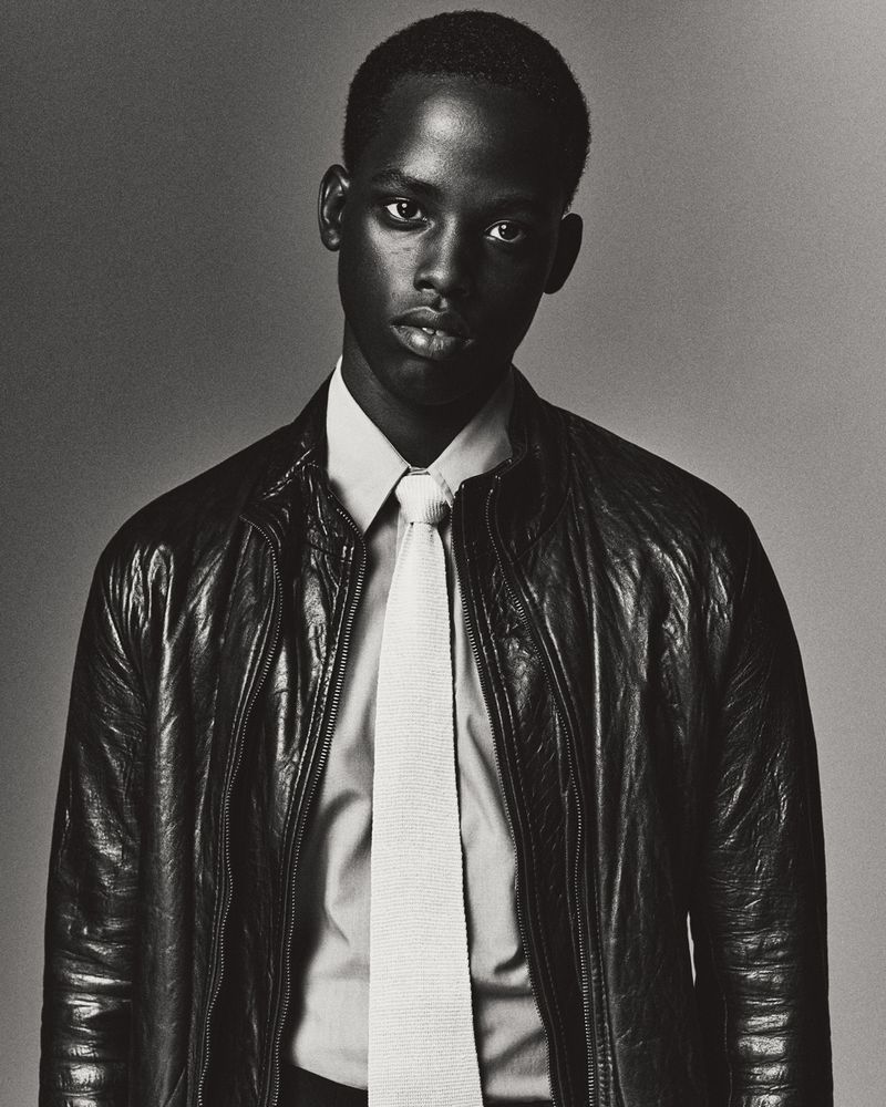 Portrait of a young black man wearing a white shirt, a light knitted tie, and a dark leather jacket against a grey background.