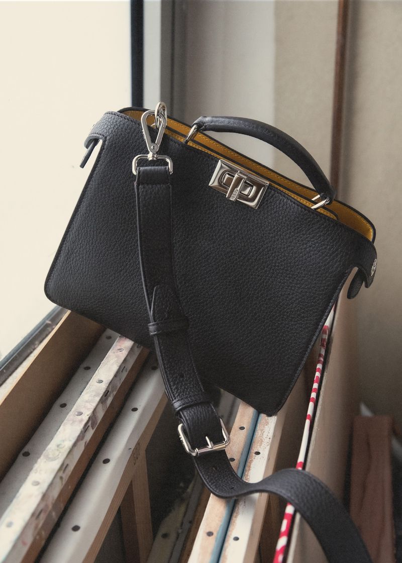 Black leather satchel handbag with top handle and shoulder strap, displayed against a window with natural light, showcasing its design.