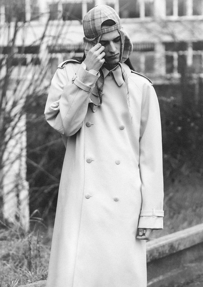 Monochrome portrait of a person wearing a long trench coat and a plaid trapper hat, standing outdoors near a building with bare trees.