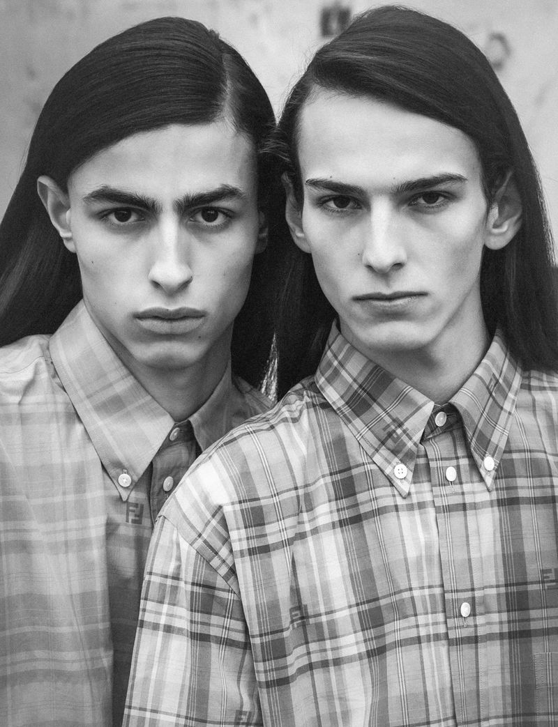 Black and white portrait of two young adult males with long hair, wearing plaid shirts, standing indoors with a serious facial expression.