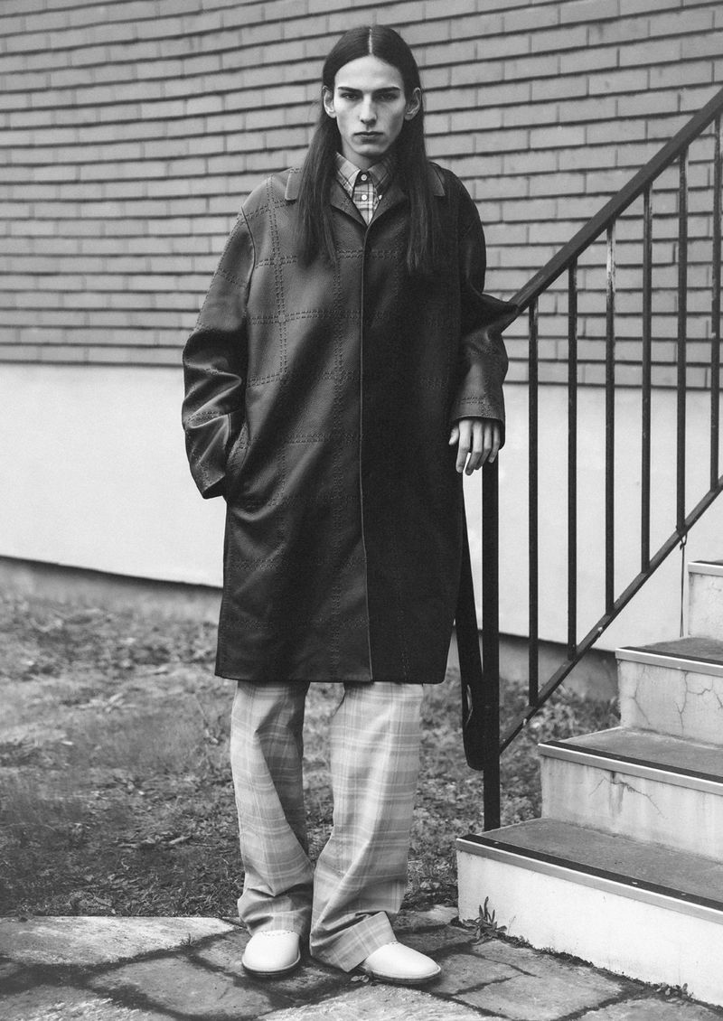 Monochrome portrait of a tall, slender adult male wearing a dark overcoat, patterned shirt, plaid pants, and white shoes. He stands by stairs.