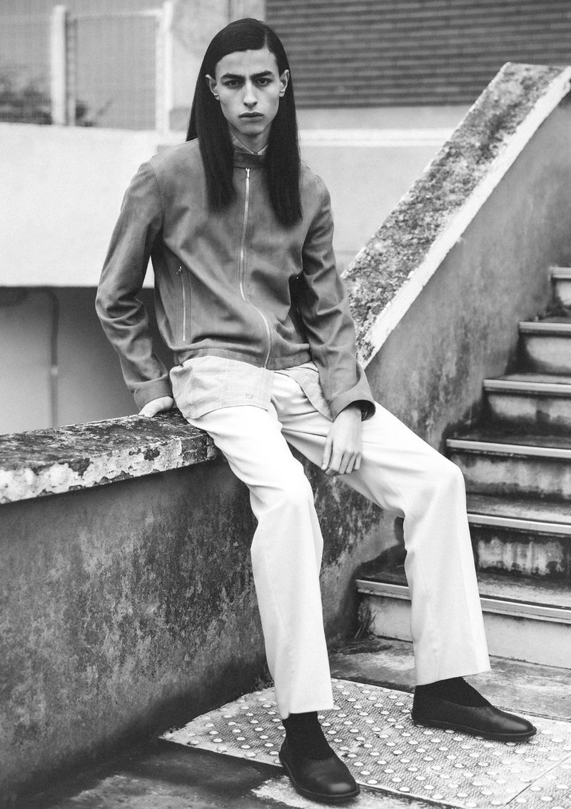 Monochrome portrait of a stylish young adult sitting on a concrete ledge wearing a jacket, pants, and shoes in a stark outdoor setting.