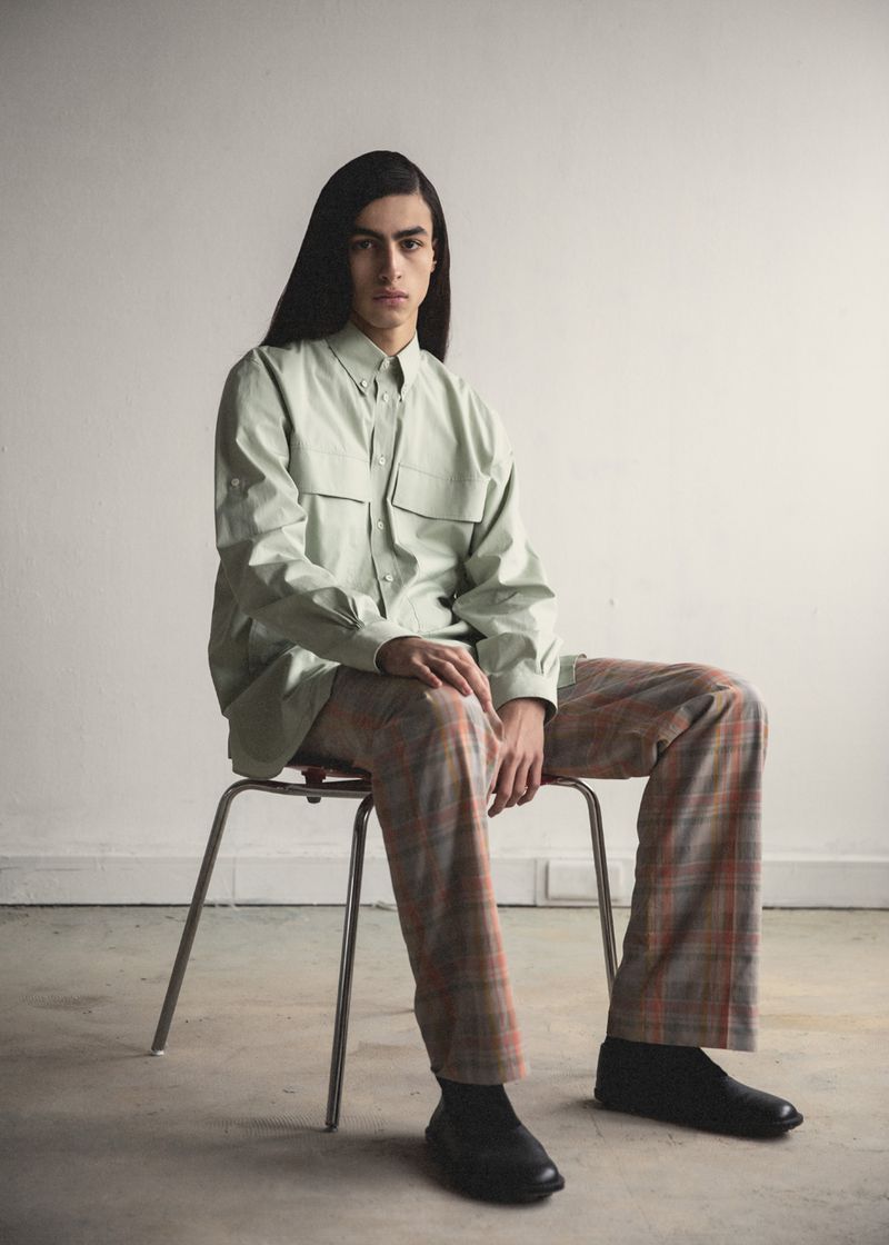 A person with long, dark hair sits on a metal chair wearing a light green button-down shirt, plaid pants, and black shoes in a studio.