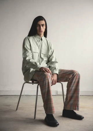 A person with long, dark hair sits on a metal chair wearing a light green button-down shirt, plaid pants, and black shoes in a studio.