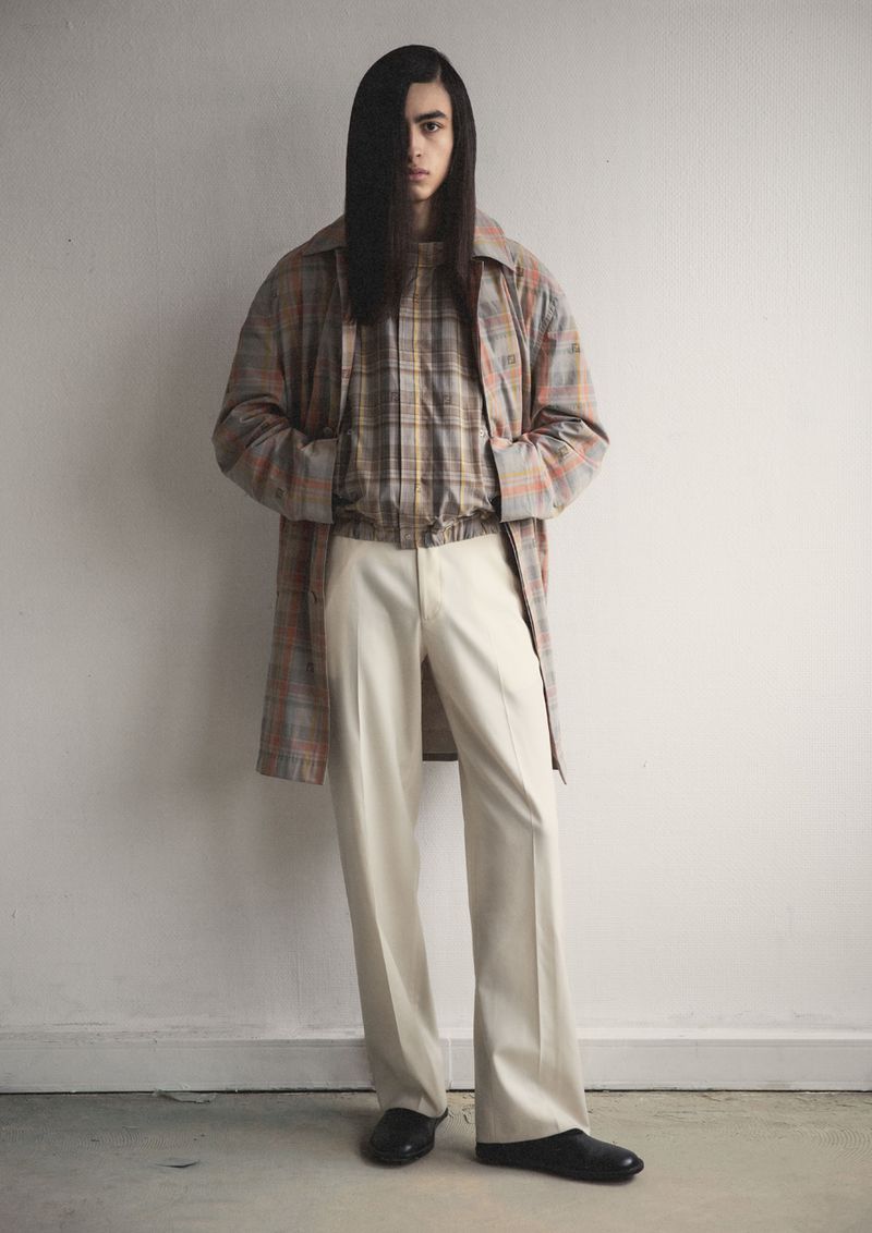 A young adult man with long dark hair poses in a studio wearing a plaid shirt and jacket with wide-leg pants and slip-on shoes.