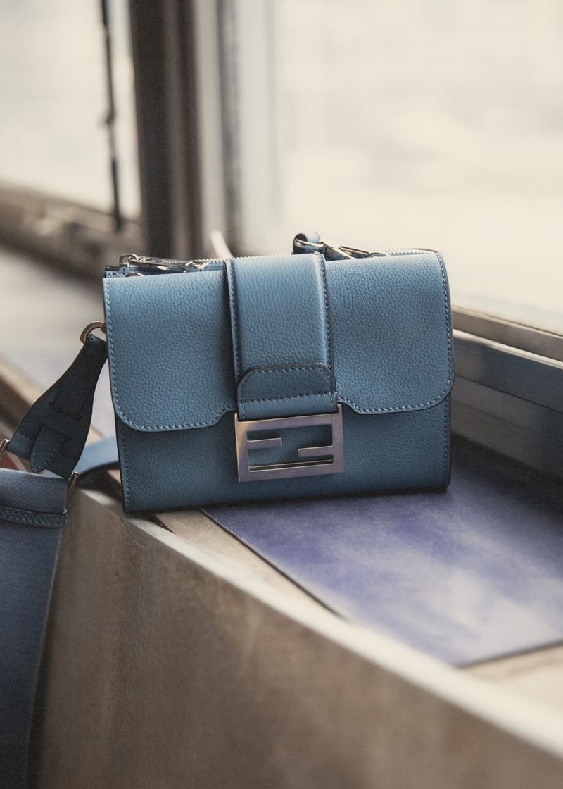 Stylish blue leather handbag with silver hardware and strap, sitting on a textured surface in a neutral indoor setting.