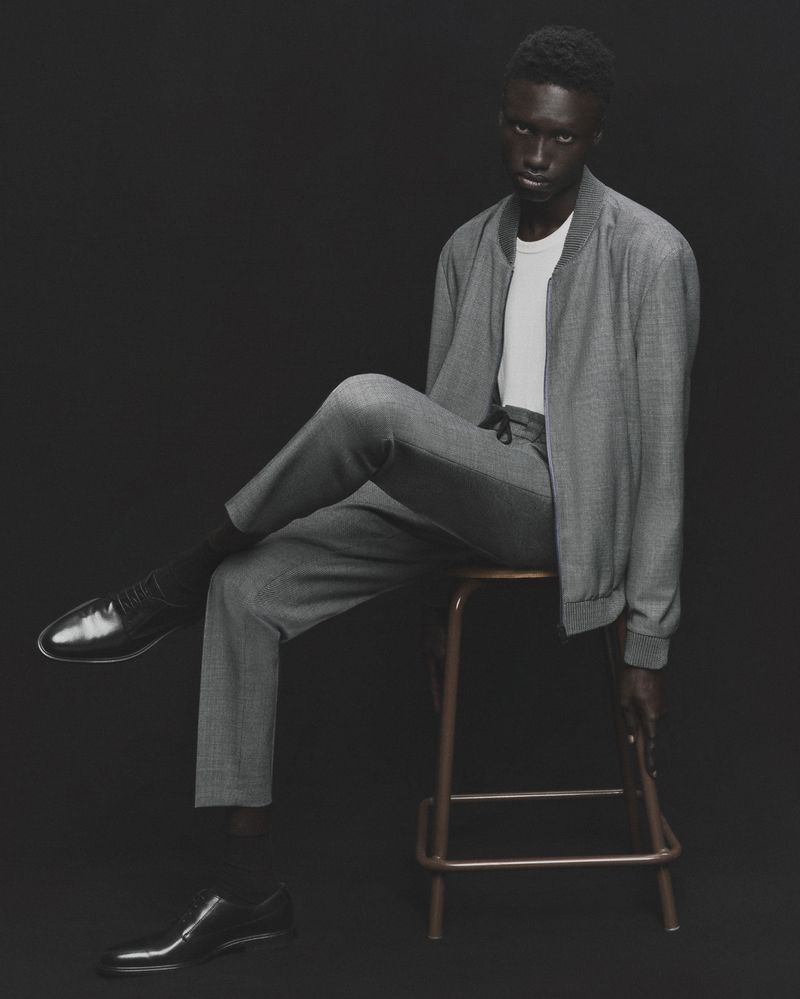 Man in a grey bomber jacket and pants sitting on a stool against a dark background, with a pair of black leather dress shoes nearby.