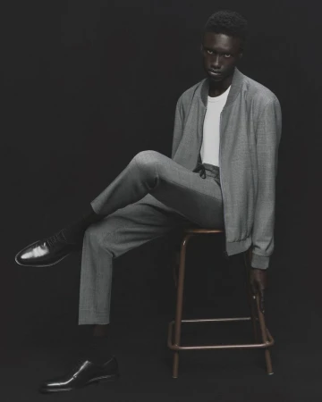 Man in a grey bomber jacket and pants sitting on a stool against a dark background, with a pair of black leather dress shoes nearby.