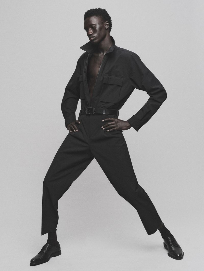 Man in black jumpsuit poses against plain backdrop. Dark-skinned male model in fashion shot showcasing modern, minimalist style.