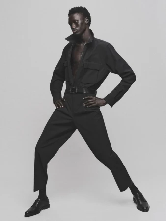 Man in black jumpsuit poses against plain backdrop. Dark-skinned male model in fashion shot showcasing modern, minimalist style.