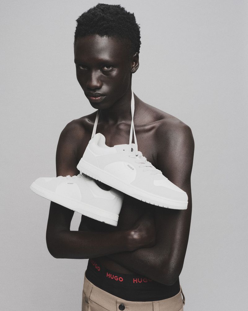 Fashion shot of a bare-chested man holding white sneakers, wearing khaki pants with visible underwear band against a grey background.