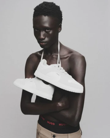 Fashion shot of a bare-chested man holding white sneakers, wearing khaki pants with visible underwear band against a grey background.