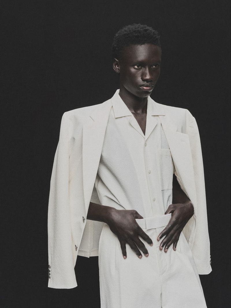 Androgynous Black model in off-white suit poses against dark backdrop in a fashion studio portrait with neutral expression.