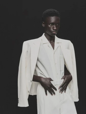 Androgynous Black model in off-white suit poses against dark backdrop in a fashion studio portrait with neutral expression.