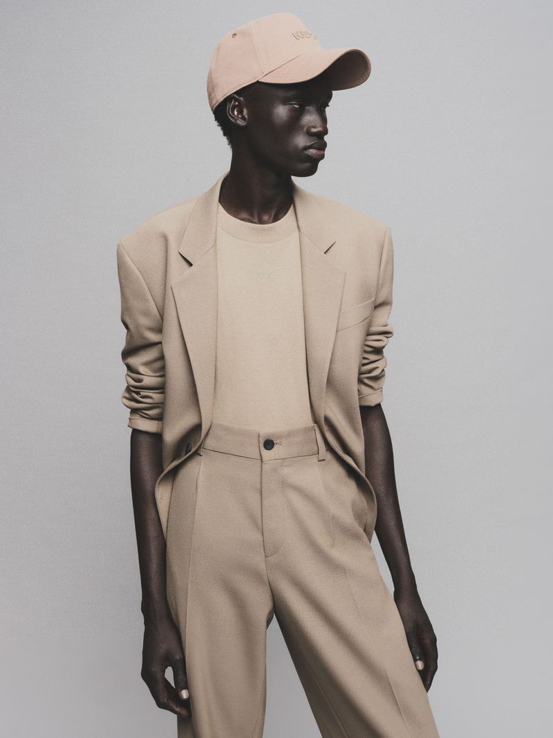 A black male model poses in a tan suit and baseball cap in a studio environment, showcasing fashion and style with a neutral background.