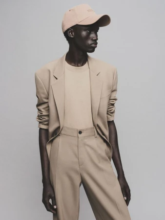A black male model poses in a tan suit and baseball cap in a studio environment, showcasing fashion and style with a neutral background.