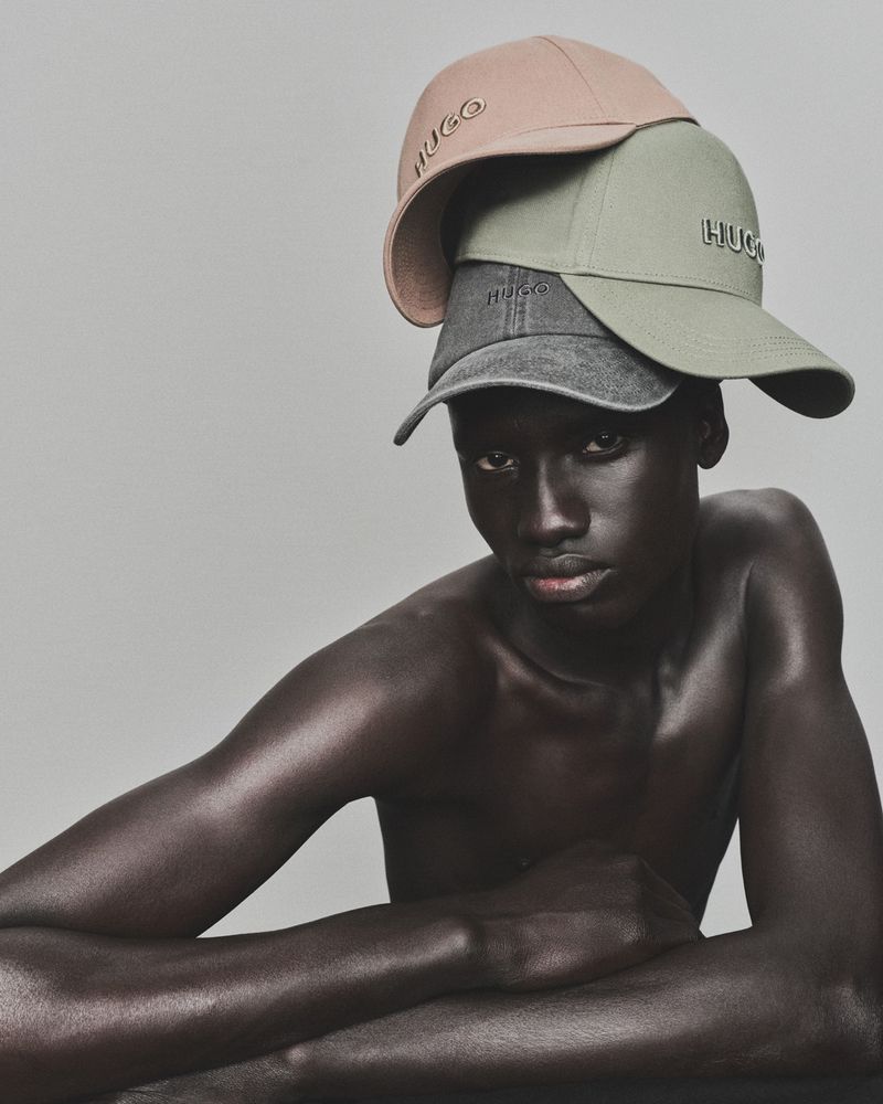Dark-skinned adult male with three layered caps from HUGO fashion label, posing in a close-up studio shot with neutral background.